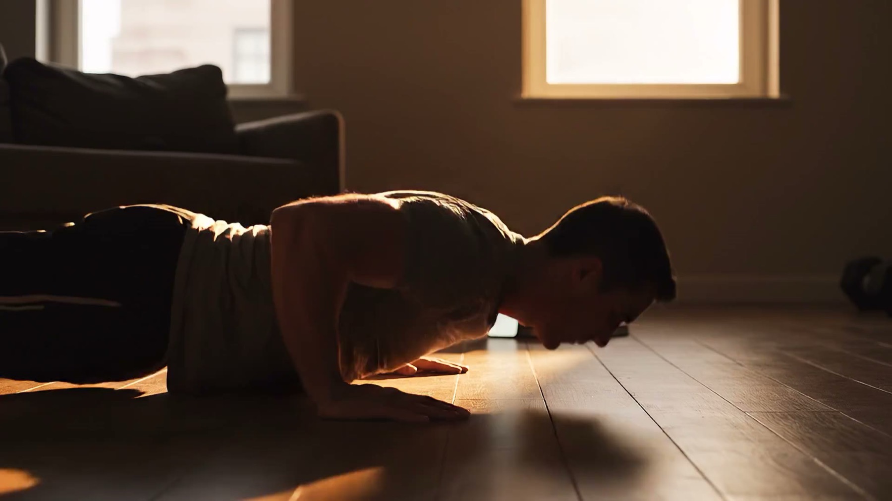 An athlete at the bottom of a push-up on a wooden floor, phone propped in front of them in low evening light.